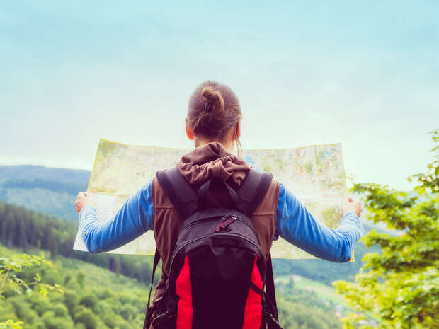 A woman stands with her back to the camera, holding a map in her hands. She wears a rucksack and is surrounded by trees.